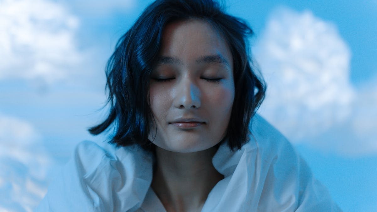 Serene portrait of a woman with closed eyes in a dreamlike, blue cloudy setting.