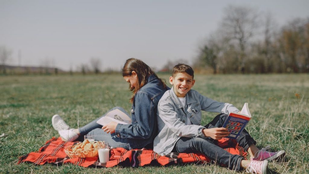 Smiling young boy in casual clothes sitting together with girl and reading interesting story while leaning back to back on checkered blanket on grass in park